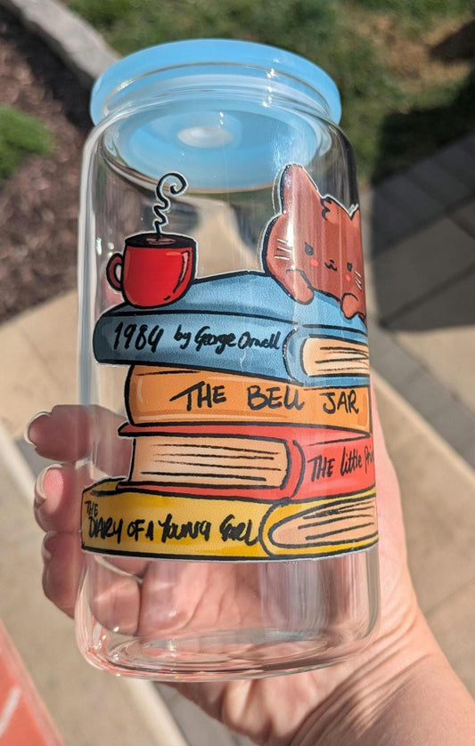 Clear tumbler with book-themed design and blue lid held by a hand.