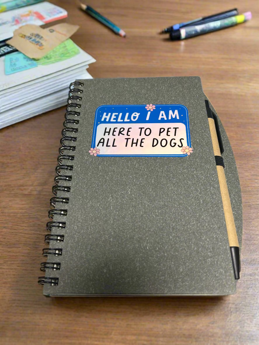 Notebook with a sticker reading 'Hello I am here to pet all the dogs' on a light-colored surface.