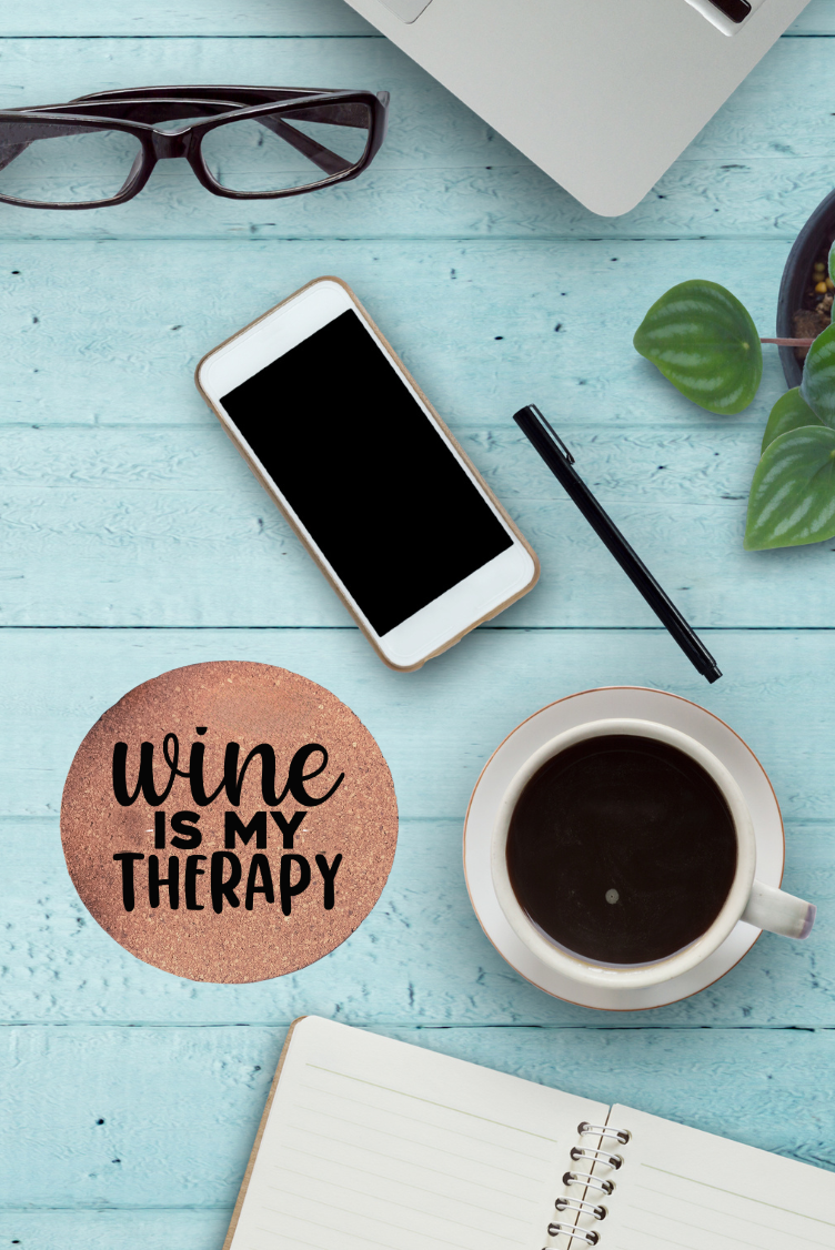 Round cork coaster with text "Wine is my therapy" on a table with a coffee cup, phone, glasses, and notebook on it.