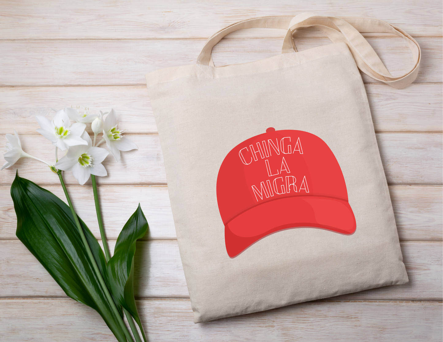 Beige tote bag with red cap graphic and text 'CHINGA LA MIGRA' on a wooden surface with white flowers.