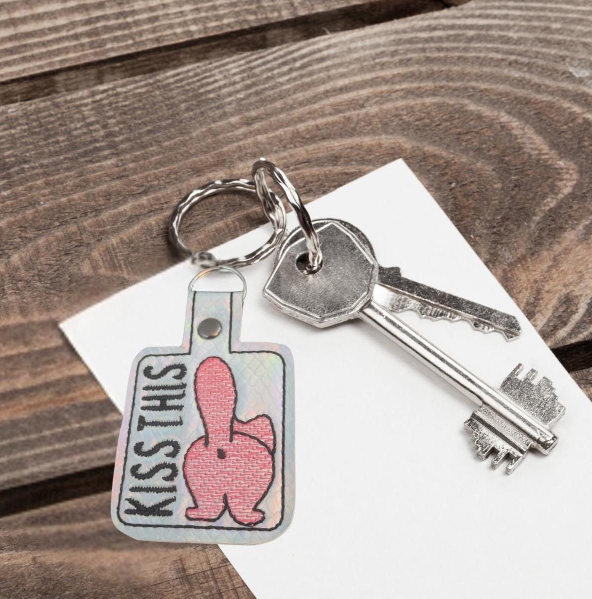 Keychain with 'Kiss This' cat butt design on a wooden surface