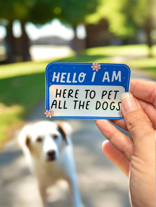 Person holding a sticker that says Hello I am here to pet all the dogs, in a sunny dog park.