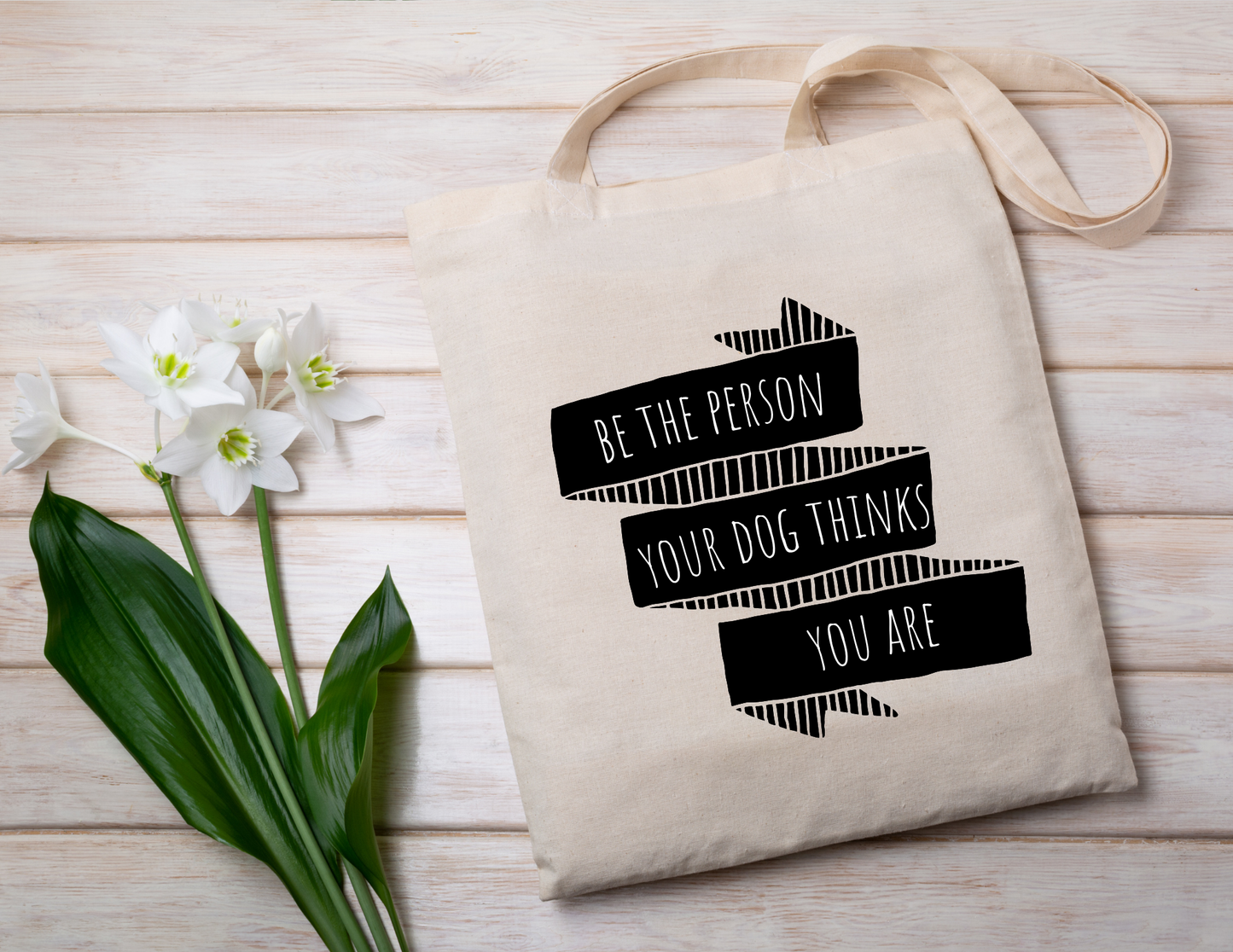 Beige tote bag with motivational quote on a wooden surface with flowers.