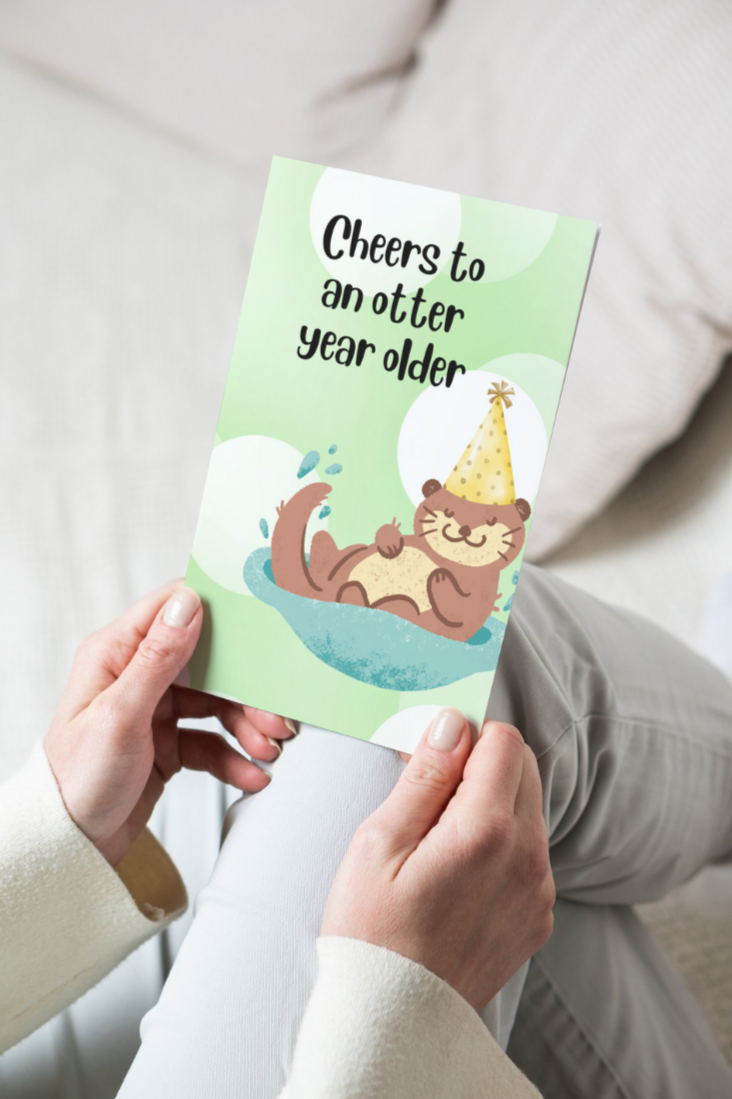 greeting card with a green and white background and an otter wearing a birthday hat, text that says cheers to an otter year older