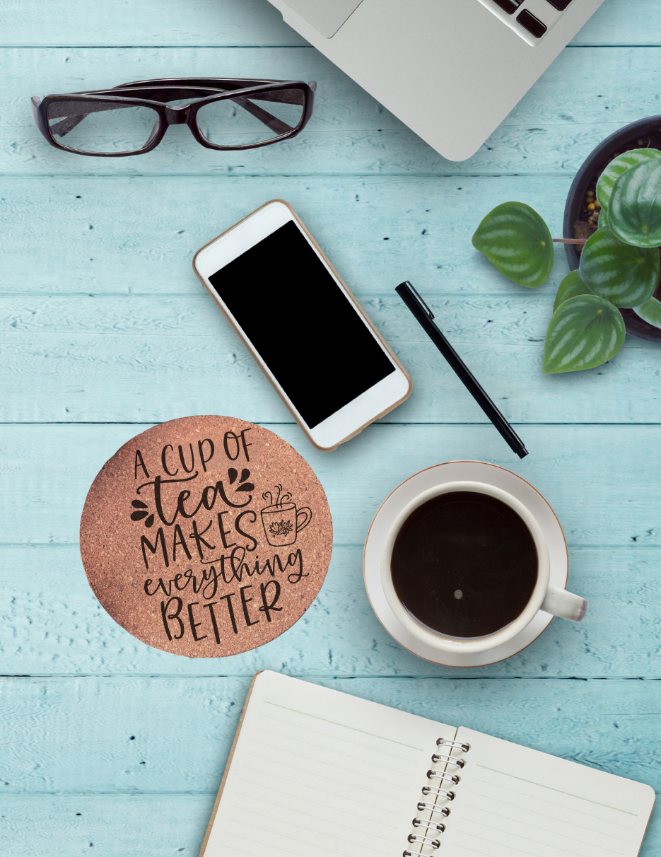 Round cork coaster with text "A cup of tea makes everything better" and a teacup graphic on a table with a coffee cup, phone, glasses, and notebook on it.