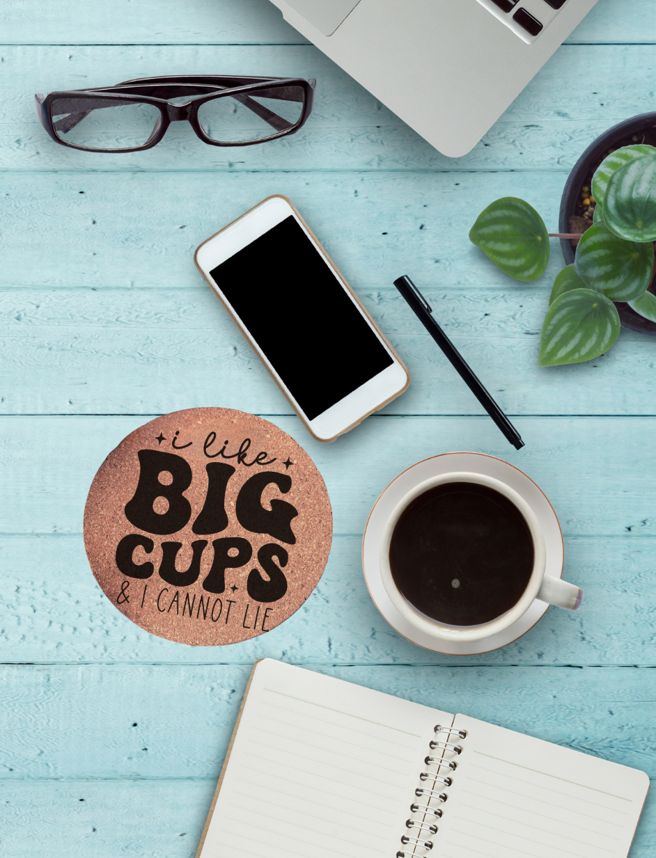 Round cork coaster with text "I like big cups & I cannot lie" on a table with a coffee cup, phone, glasses, and notebook on it.