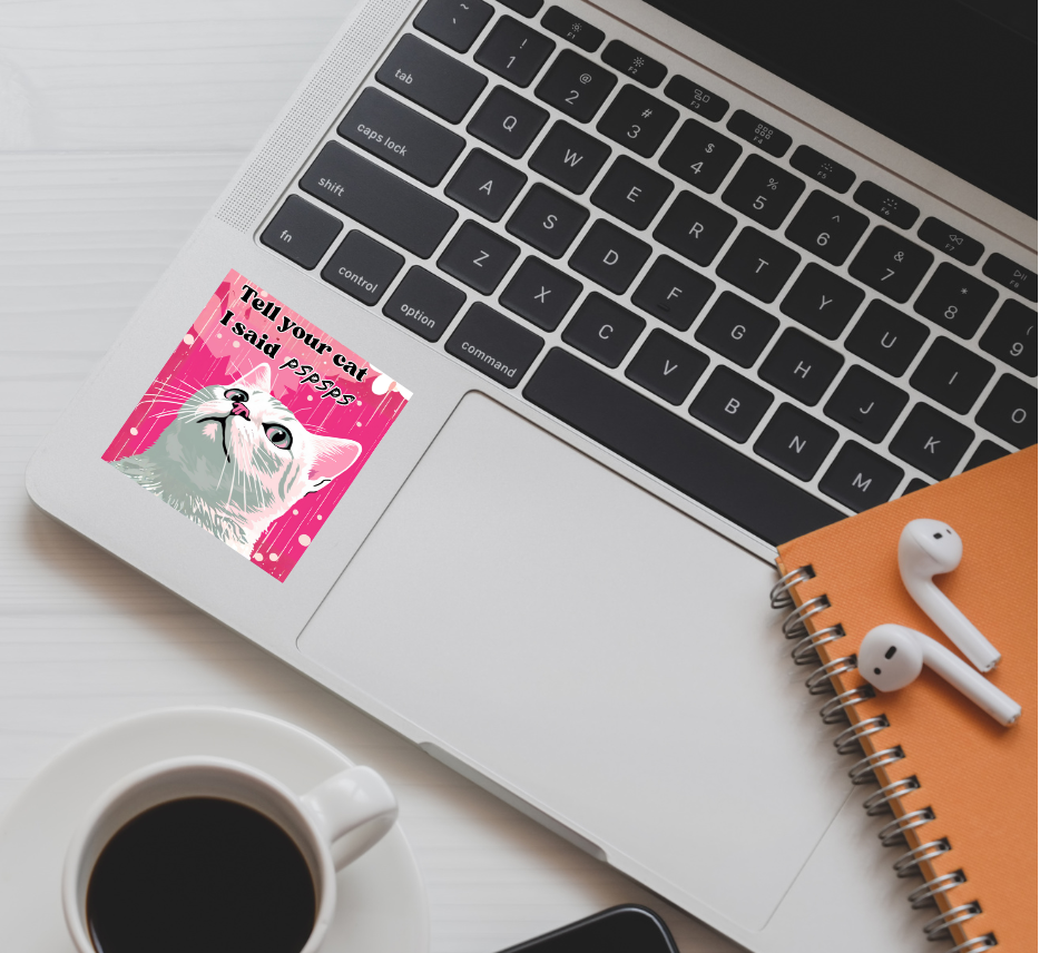 Laptop with a tell your cat I said pspsps sticker on the palm rest area, next to a coffee cup and earphones on a white surface.