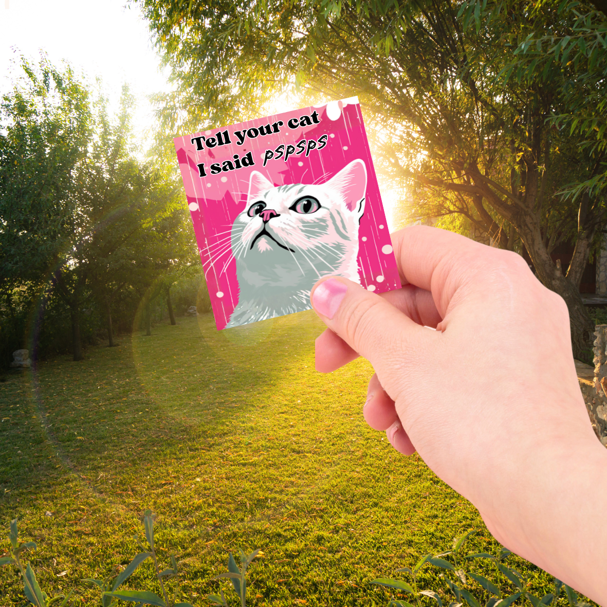 Hand holding a card with a cat illustration and "Tell your cat I said pspsps" text in a park setting