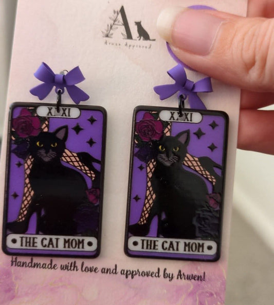 Pair of 'The Cat Mom' earrings on a purple card held by a hand.