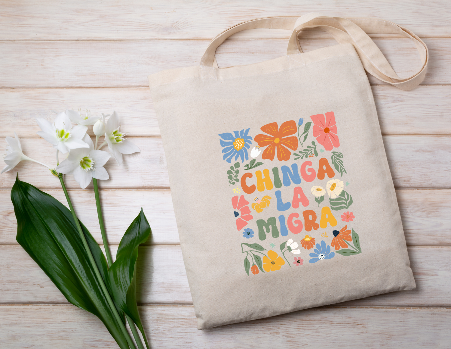 Beige tote bag with colorful floral design and "Chinga la migra" text on a wooden surface with flowers.