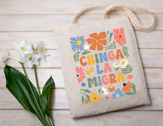 Tote bag with colorful floral design and "chinga la migra" text on a wooden surface with flowers.