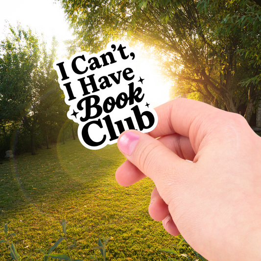 Hand holding a 'I Can't, I Have Book Club' sticker with a scenic background of trees and sunlight.