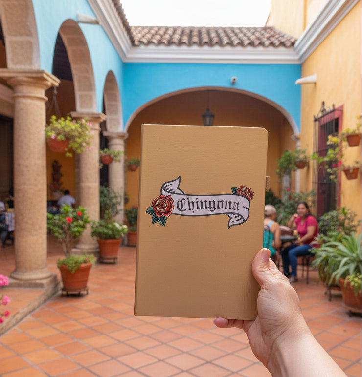 Hand holding a gold notebook with 'Chingona' design in a courtyard