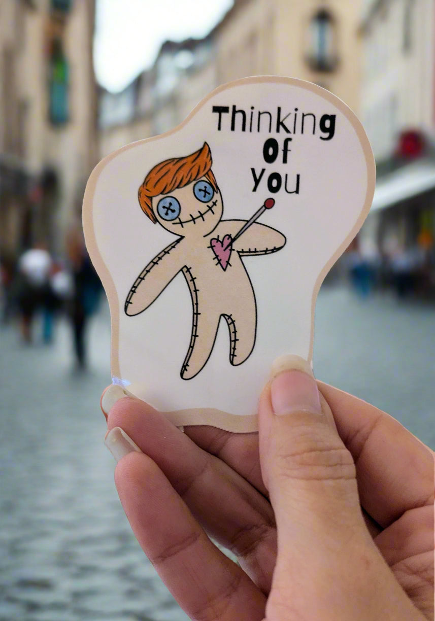 Hand holding a sticker with a voodoo doll illustration of a man with orange hair and 'Thinking of you' text.