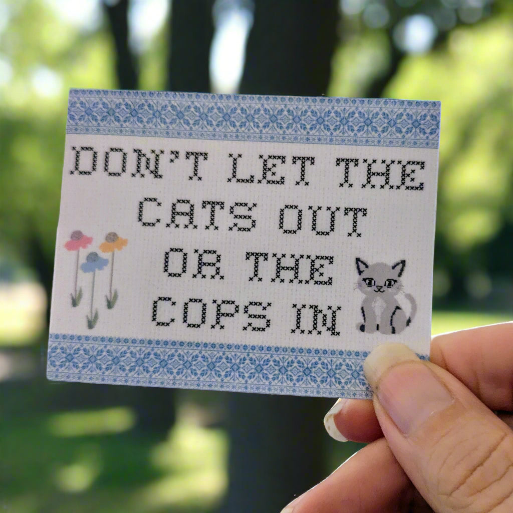 Hand holding a cross-stitched looking sticker with text and a cat illustration against a blurred green outdoor background.