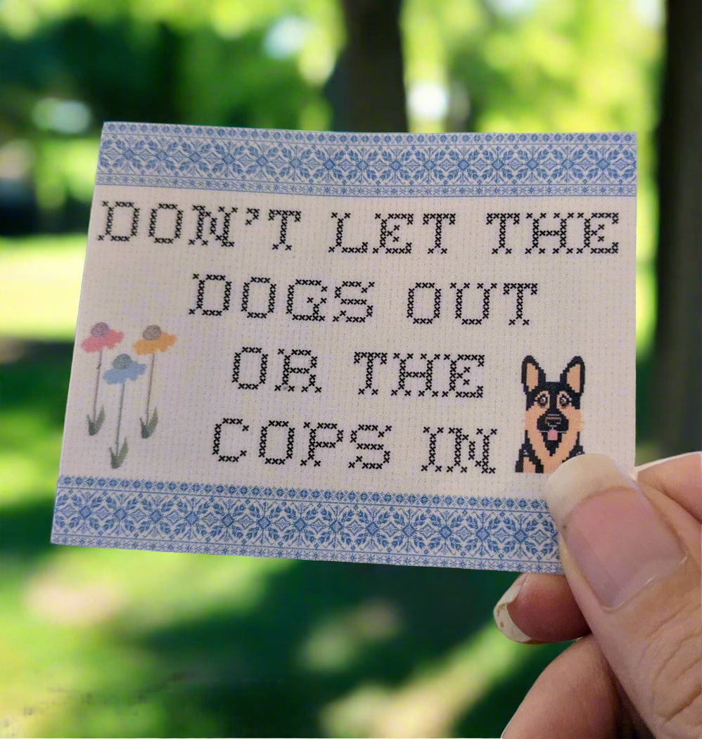 Cross-stitched looking sticker with text 'Don't let the dogs out or the cops in' held by a hand.