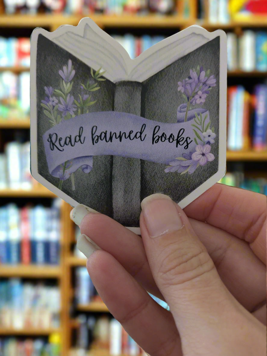 Hand holding a small book-shaped stickerwith lavender flowers and 'Read banned books' text.