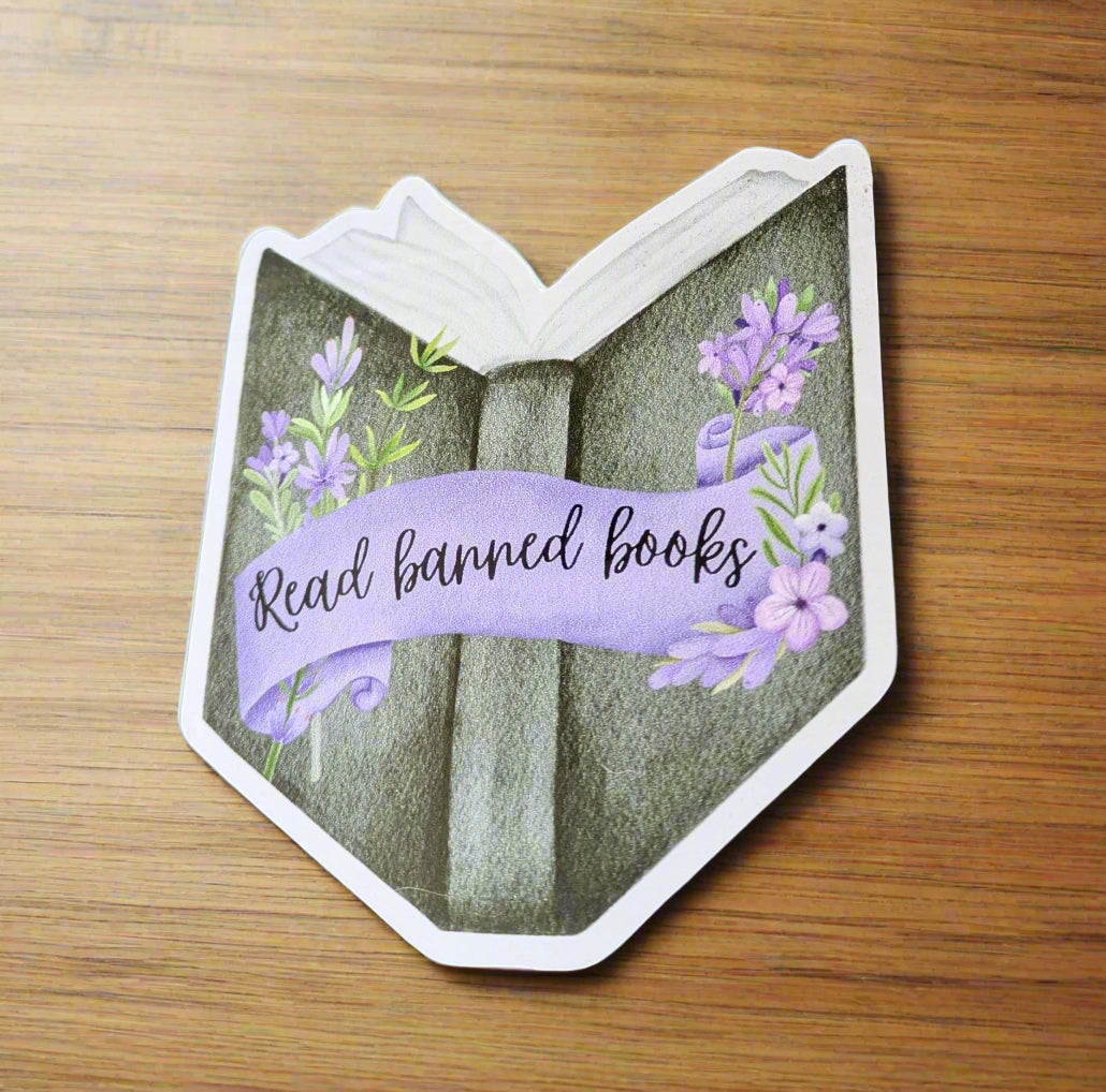 Decorative book-shaped sticker with lavender flowers and 'Read banned books' text on a wooden surface.