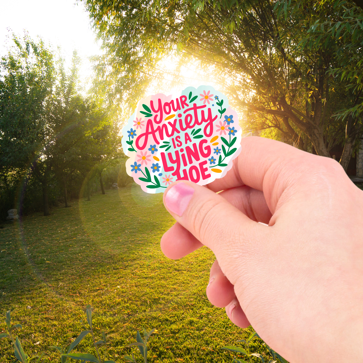 Hand holding a colorful sticker with "Your anxiety is a lying hoe" text in a park setting