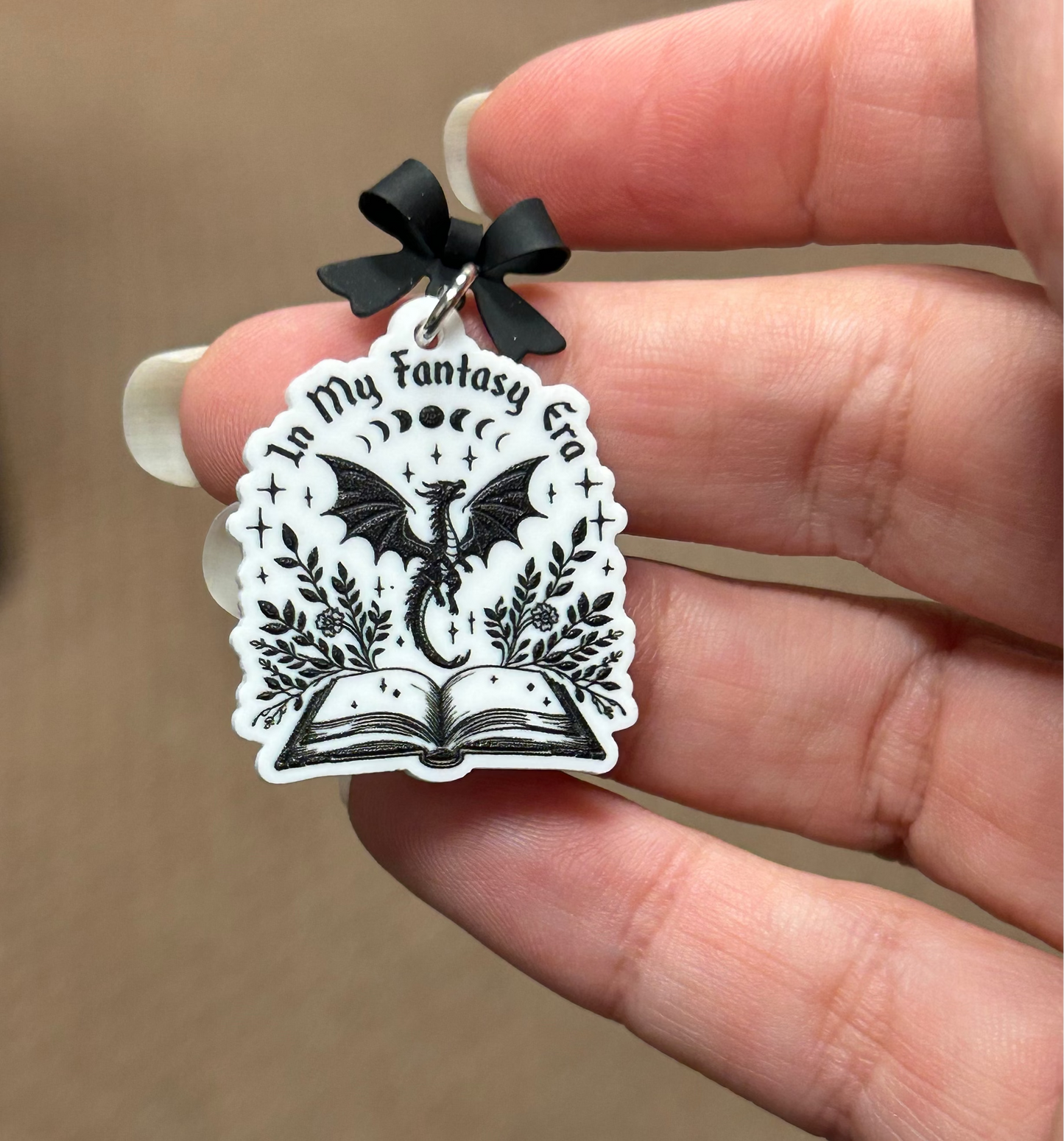 Hand holding a bow stud earring with a dragon graphic and "in my fantasy era" text.