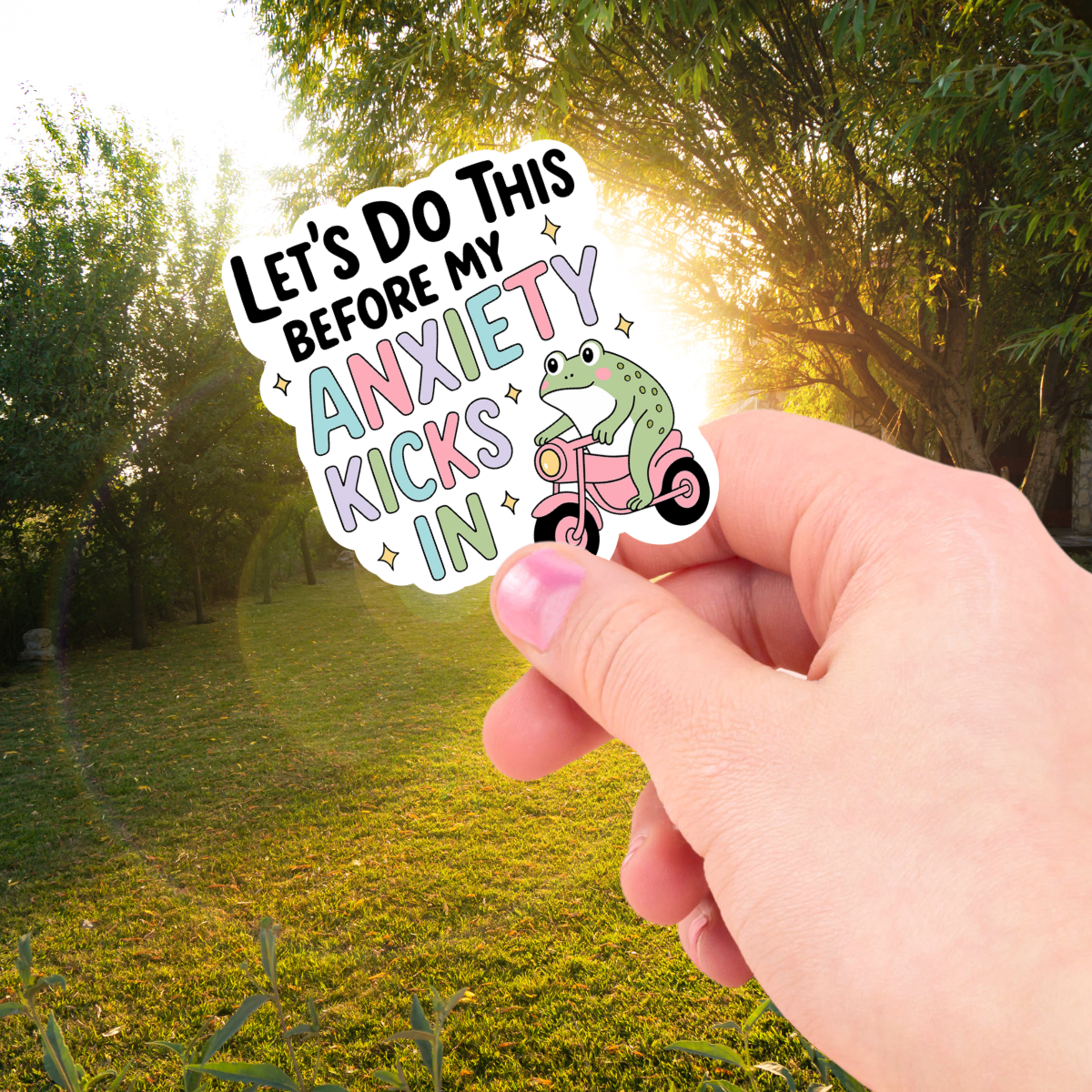 Hand holding a sticker with a frog on a scooter and "let's do this before my anxiety kicks in" motivational message against a natural background