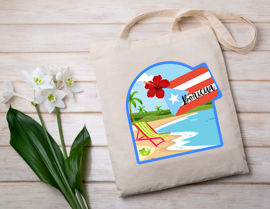 Beige tote bag with a beach scene design featuring 'Boricua' and a Puerto Rican flag on a wooden surface with flowers.