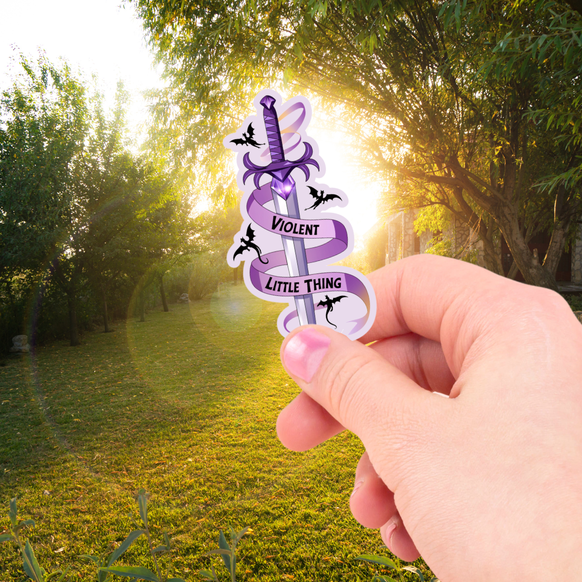 Hand holding a purple and white sticker with "violent little thing" text and a sword design against a natural background.
