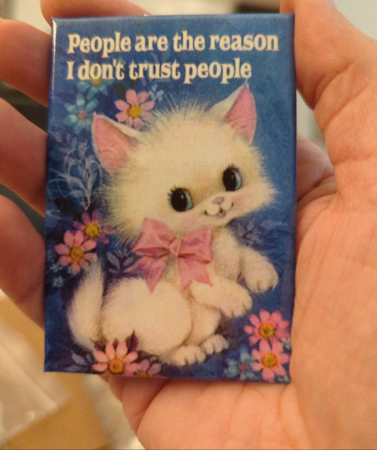 Fridge magnet with a cartoon cat and text 'People are the reason I don't trust people' held in a hand.