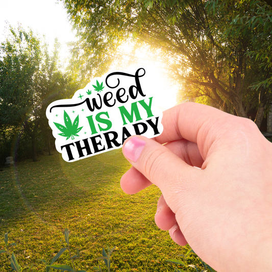 Hand holding a 'weed is my therapy' sticker with a nature background